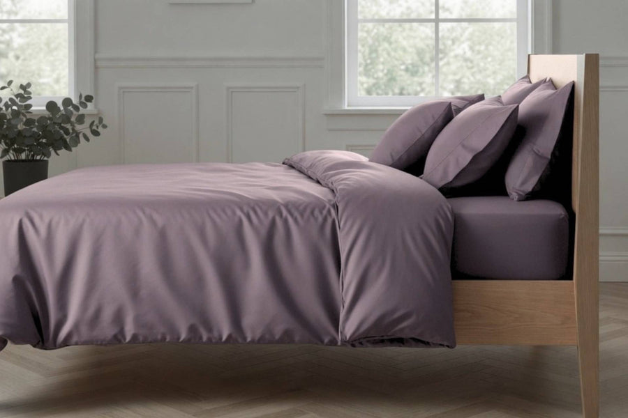 Bed with purple bedding and pillows in a room with large windows and plants.