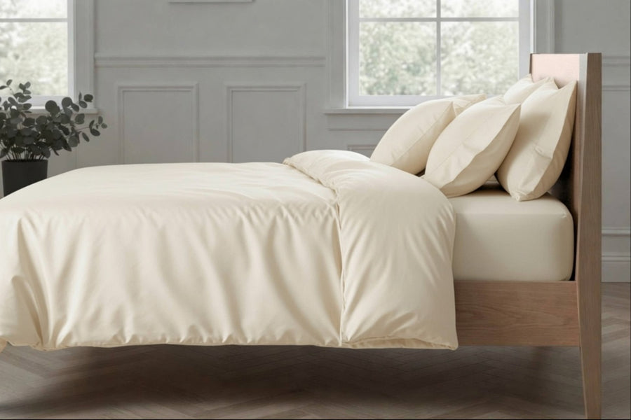 A neatly made bed with cream-colored bedding, including a duvet cover and four matching pillows, set against a light gray paneled wall with a window and a wooden headboard.