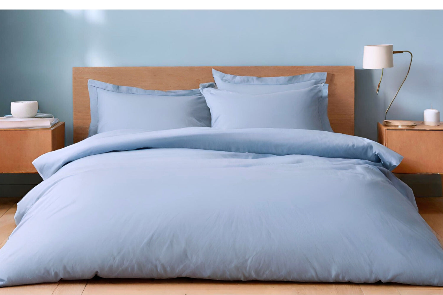 A duvet cover in a light powder blue color, displayed on a bed in a room with a wooden headboard and a bedside table with a lamp. House of March bed linens