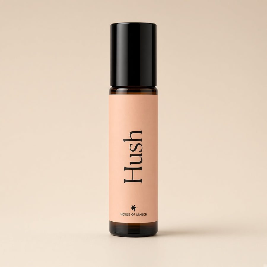 A small amber glass roller bottle with a black cap and a peach-pink label reading 'Hush' and 'HOUSE OF MARSH', centered on a warm beige background.